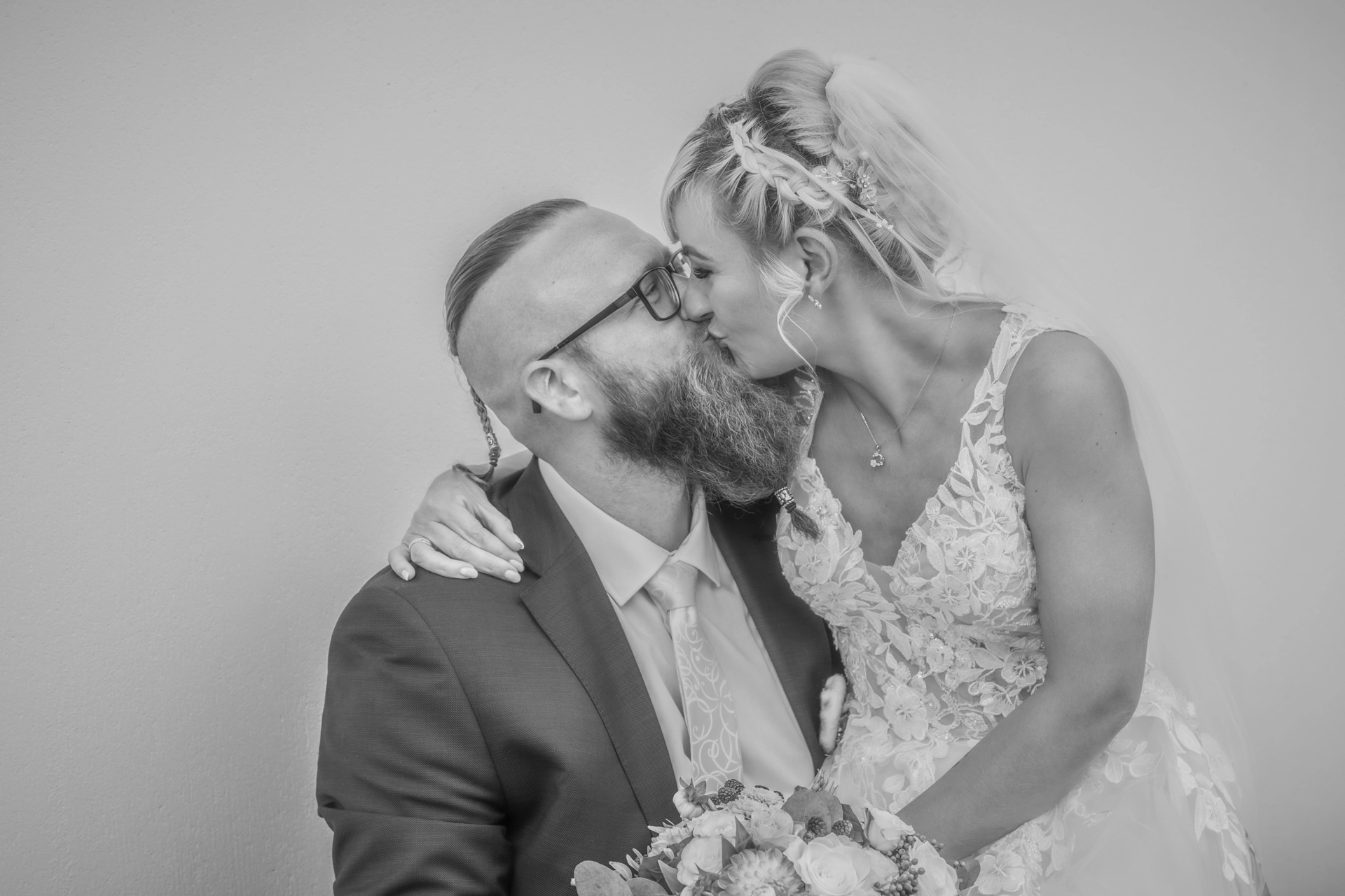 Zeitlose Liebe in Schwarz-Weiß Schwarz-weiß Hochzeitsfoto: Glückliches Brautpaar küsst sich innig, Bräutigam mit Bart und Undercut, Braut im Spitzenbrautkleid mit Blumenstrauß. Emotionale Hochzeitsfotografie in klassischem Stil."